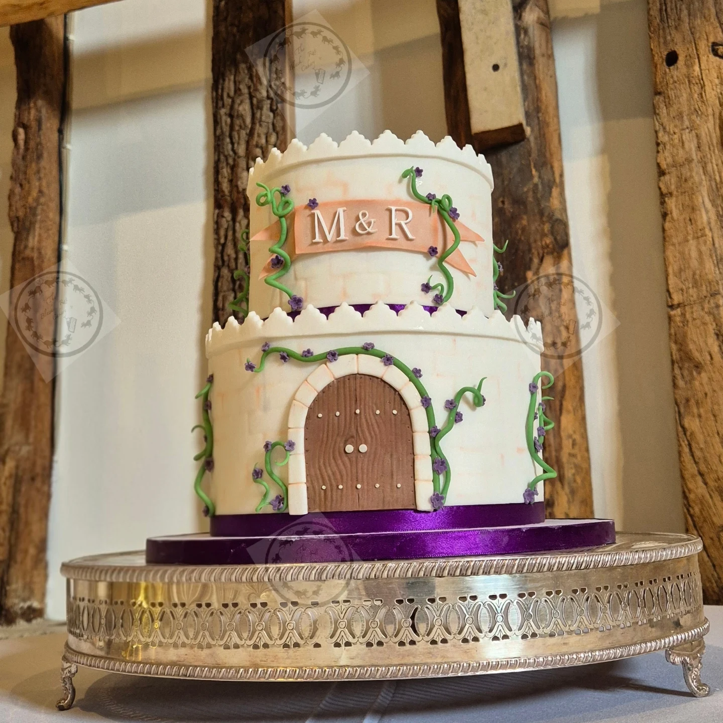 Two-tier ivory wedding cake styled as a miniature castle with a fondant door, climbing vines and a personalised initial banner